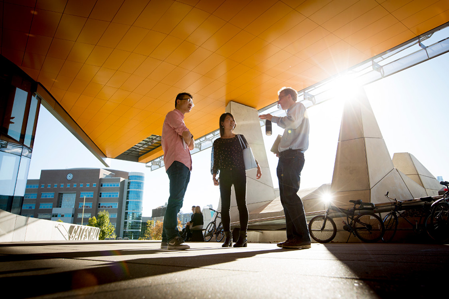 Three people talking outside a modern building with bikes and sunlight behind them.